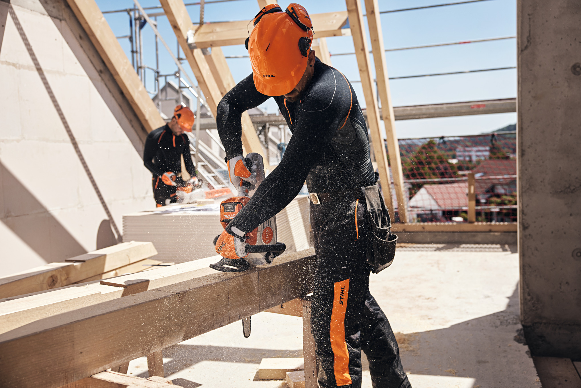 Profesjonalista STIHL na więźbie dachowej przecina drewnianą belkę akumulatorową pilarką spalinową, podczas gdy w tle inny pracownik używa drugiej akumulatorowej pilarki