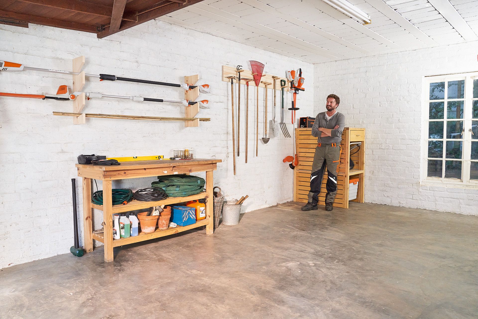 Mężczyzna ze skrzyżowanymi ramionami opiera się obok samodzielnie wykonanego uchwytu na narzędzia ogrodowe w garażu 