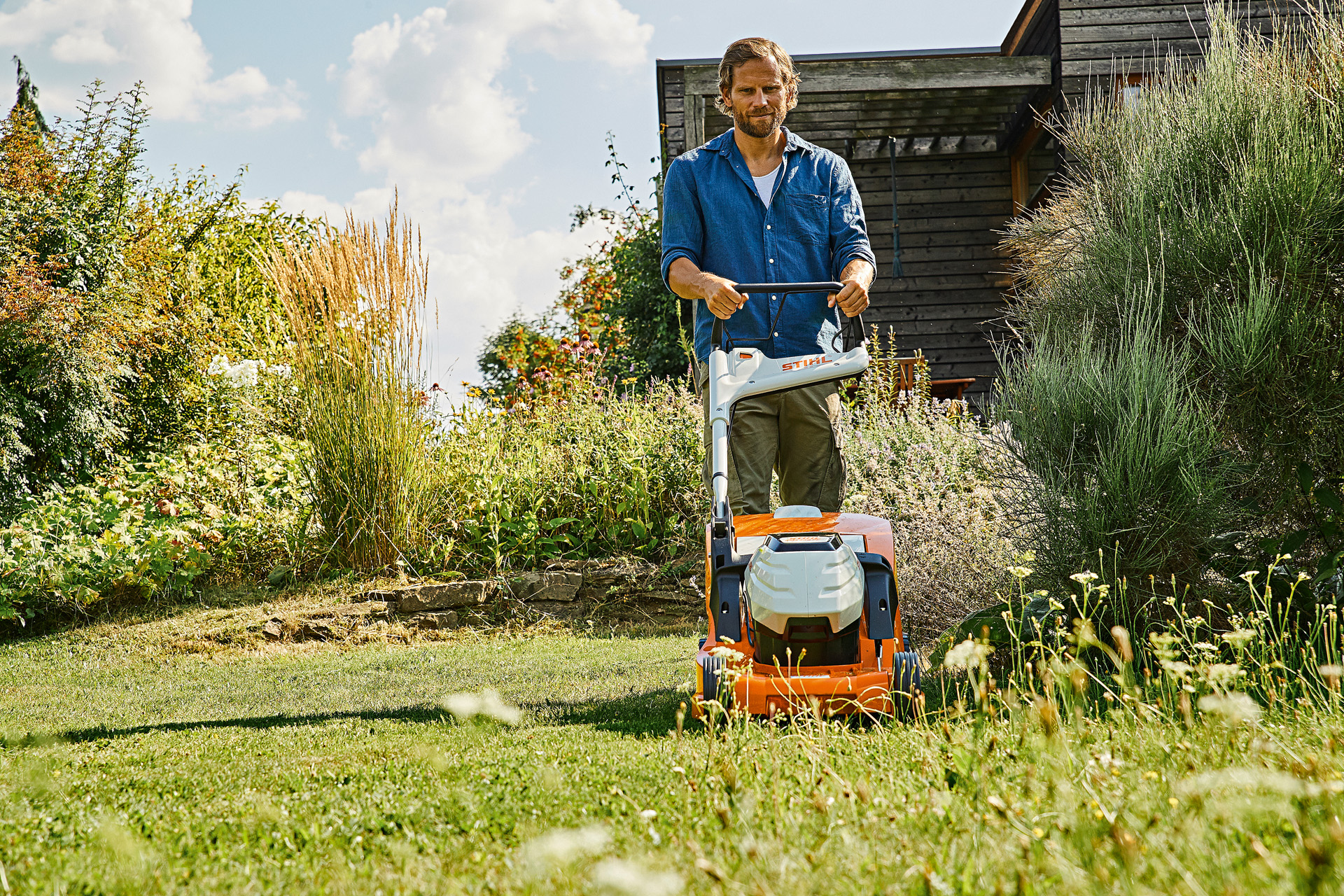  Mężczyzna jest w trakcie koszenia zaniedbanej i zarośniętej trawy akumulatorową kosiarką STIHL RMA 448 P-C w ogrodzie obsadzonym wysokimi krzewami i roślinami.